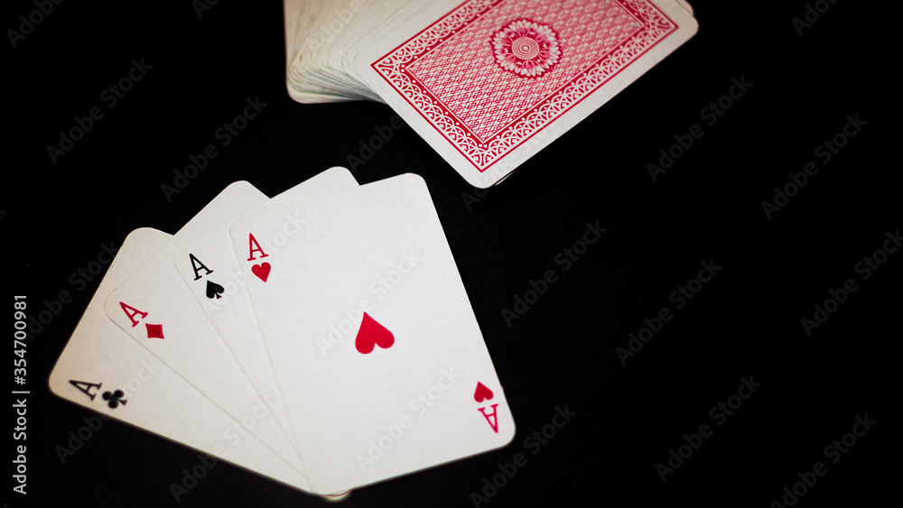 Obraz premium Ángulo levemente elevado de un juego de ases de poker aislados sobre un fondo negro y con iluminación directa sobre ellos. Junto se encuentra un mazo de cartas de fondo rojo. 