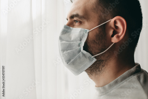 Young brunette white european man at home on quarantine with medical mask near window white backround. Coronavirus, illness, infection, quarantine, surgical bandage. europe, medical mask, depression