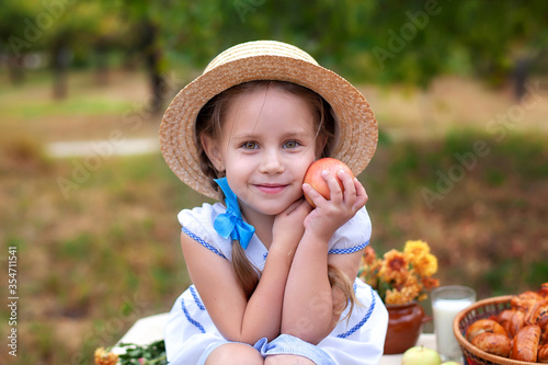 Little girl in straw hat picking apples in green grass background. Child Girl with red apple in summer orchard. Harvest concept. Smiling child on picnic holding an apple in garden. Gardening. 