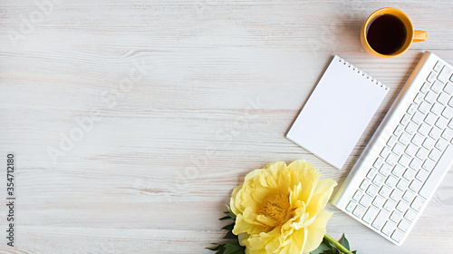 Banner with copy space - a blank notebook surrounded by yellow peony flower, coffee cup and keyboard, a light wooden background, To-do list, plans, dreams, work from home. Top view,