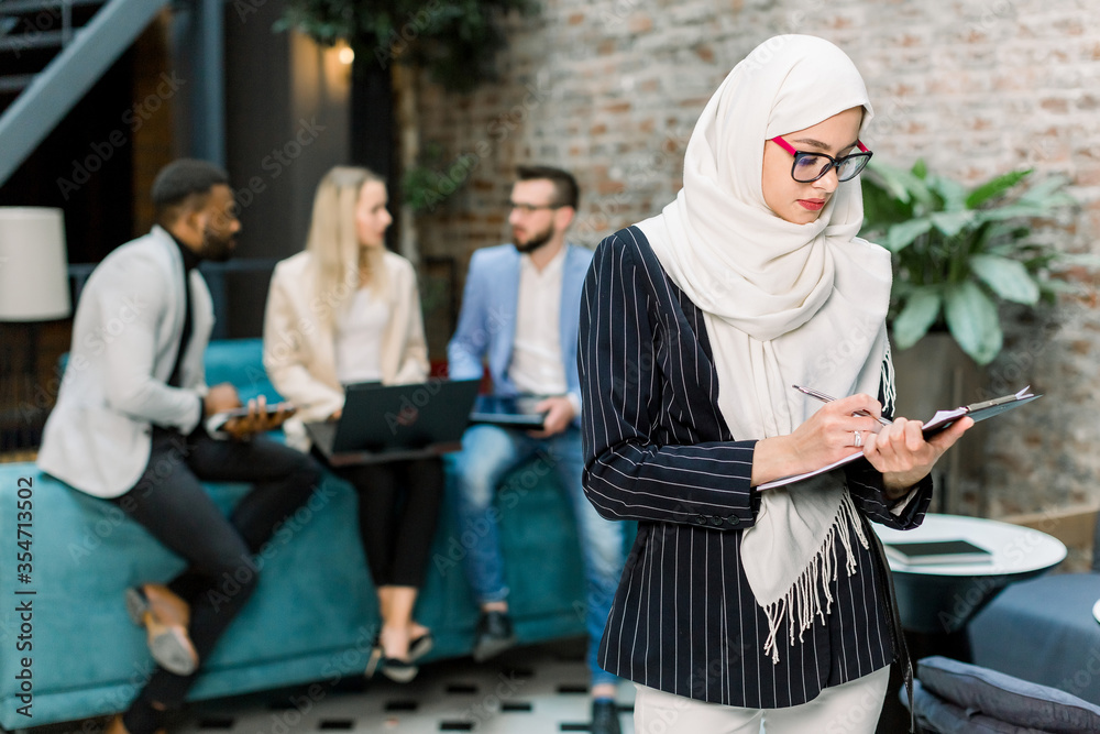 Young pretty Muslim business woman wearing white hijab signing contract or financial papers standing in modern office. Three multiethnic office colleagues on the background working together