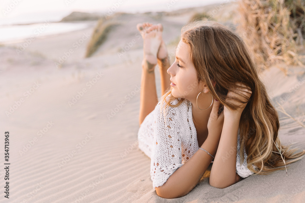 Portrait of happy young woman in white dress, lies on the sand dunes next to the sea. in summer time, on the sunset. Portrait of happy young woman in white dress, lies on the sand dunes next to the sea. in summer time, on the sunset.