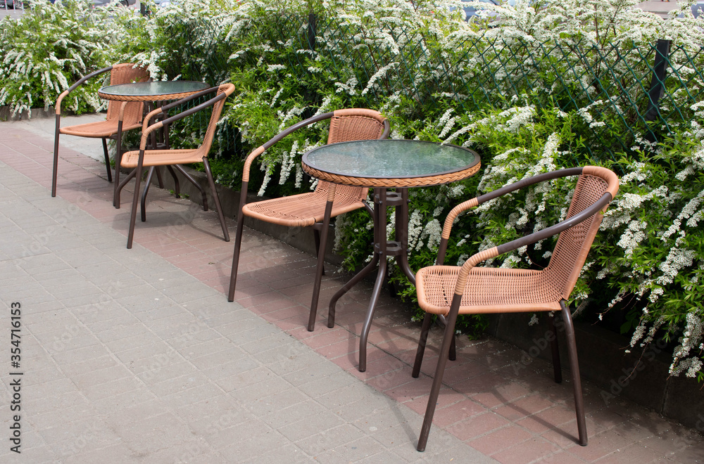 Summer street cafe. The glass table and brown chairs are outdoors. A small cozy restaurant with retro furniture and bushes with white flowers.