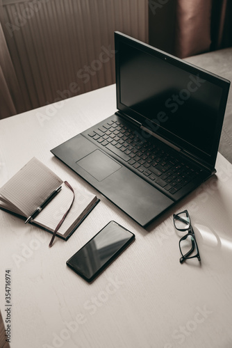 Business workplace, man working at laptop, writing on notebook with pen, glasses and smartphone on white table 