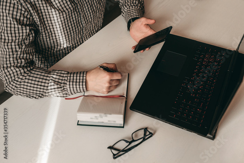 Business workplace, man working at black laptop, writing on notebook with pen, black glasses and holding smartphone on white wooden table