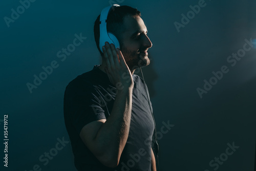 Young bearded man in glasses with headphones listens to music on dark blue background in nightclub. © lesia17