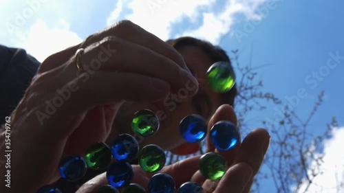 High quality close-up 4k slow motion video of a man holding blue and green glass marbles and putting balls on a glass top table, view from the bottom, blue sky abstract background