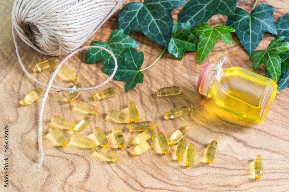 Fototapeta premium Transparent gelatinous oblong capsules of fish oil were scattered on the wooden background near the oil bottle. Omega-3-essential polyunsaturated fatty acids. The neck of the bottle is tied with rope