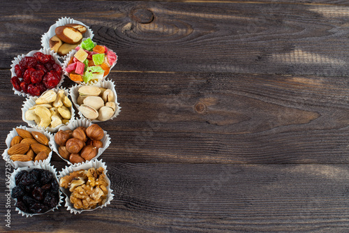 A serving of assorted candied fruit, dried cherries, almonds, raisins, walnuts and hazelnuts in paper muffin cups on a dark wood background. Copyspace.