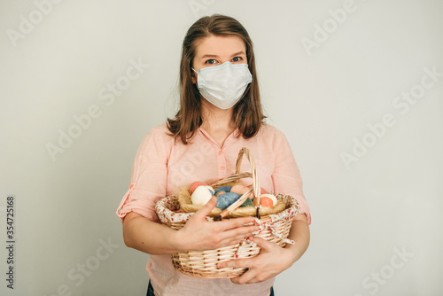 young woman with Easter at the basket and colored eggs she holds in her hands on white backround. quarantine at home in medical surgical masks. Basket, eggs, holiday, coronavirus, illness, stay