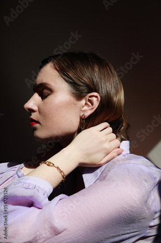 Side portrait of attractive young woman standing against dark background wearing lavender shirt. Vertical shot.