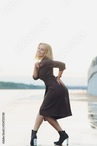 Outdoor portrait of young beautiful caucasuan woman with long straight hair in short black dress, beige shoes holding brown leather handbag walking city street. photo