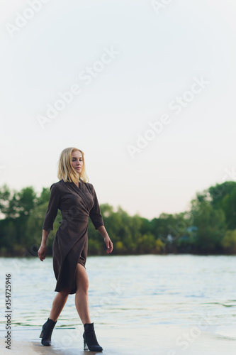 Outdoor portrait of young beautiful caucasuan woman with long straight hair in short black dress, beige shoes holding brown leather handbag walking city street. photo