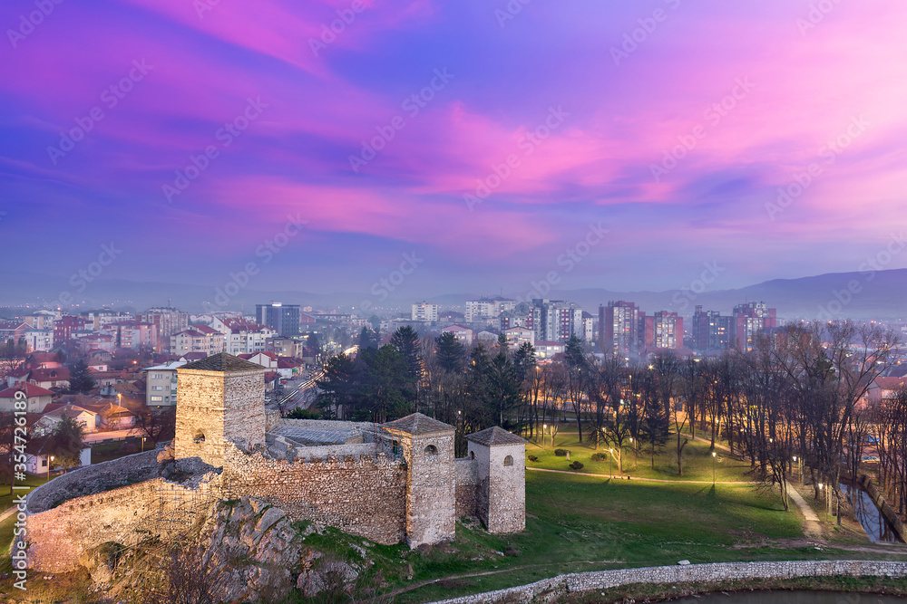 Colorful, purple sunset and fluffy, sunlit magenta clouds over blue hour Pirot cityscape and a foreground ancient fortress