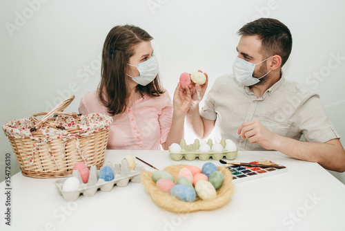 Young white European couple celebrating paint Easter quarantine at home in medical surgical masks. Basket, eggs, holiday, coronavirus, illness, stay home, infection, flue, surgical bandage.
