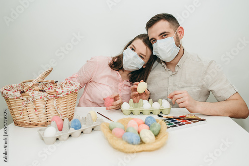 Young white European couple celebrating paint Easter quarantine at home in medical surgical masks. Basket, eggs, holiday, coronavirus, illness, stay home, infection, flue, surgical bandage.