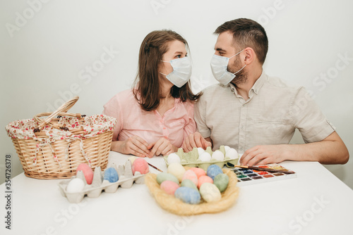 Young white European couple celebrating paint Easter quarantine at home in medical surgical masks. Basket, eggs, holiday, coronavirus, illness, stay home, infection, flue, surgical bandage.