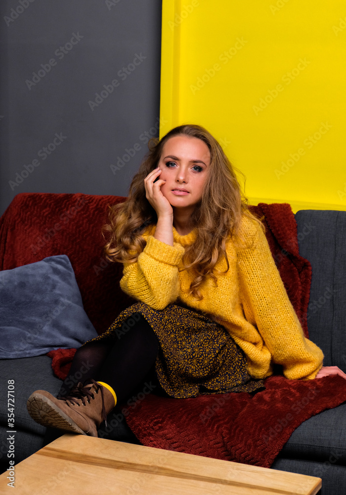 Portrait of girl in yellow oversize sweater, skirt and brown boots. Woman sitting on sofa. Yellow square on the background. Portrait of girl in yellow oversize sweater, skirt and brown boots. Woman sitting on sofa. Yellow square on the background.