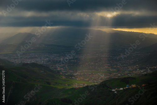 The rays of the sun make their way through the dark clouds on the green hills with houses in the Tenerife, Canary island