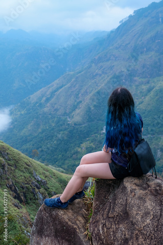 A young girl with a long blue hair and a black backpack sitting on a cliff. Famous mountain in southern Sri Lanka known as Little Adam's Peak. Lonely girl in isolated place. Hiking places in Asia.