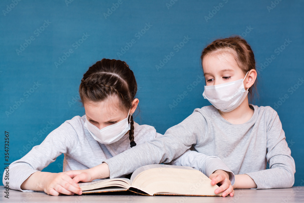 A girl in a medical mask in quarantine reads a book, and another girl in a mask takes it from her. Education for children in isolation in an epidemic
