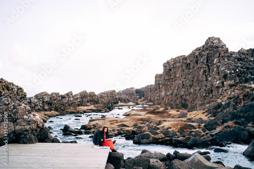 Fototapeta Naklejka Na Ścianę i Meble -  A guy and a girl are sitting embracing on a bridge near Ehsaraurfoss Waterfall, Ehsarau River, National Park, Thingvedlir, the region of Sudurland, Iceland.