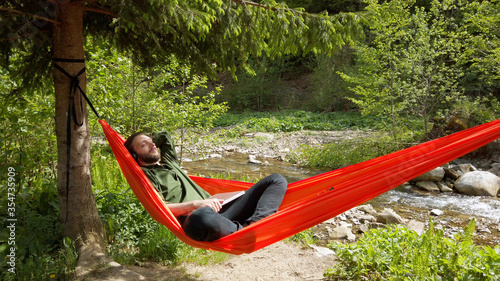 Relaxed man lying in hammock outdoor and listening music with wireless headphones. Enjoying nature while quarantine