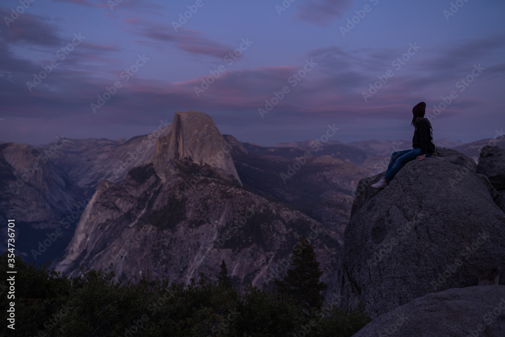 A young girl in blue jeans, white sneakers and a dark fable and hat sits on a large rock and looks at the endless landscape of mountains, evening cloudy sky and forests in the valley of California USA