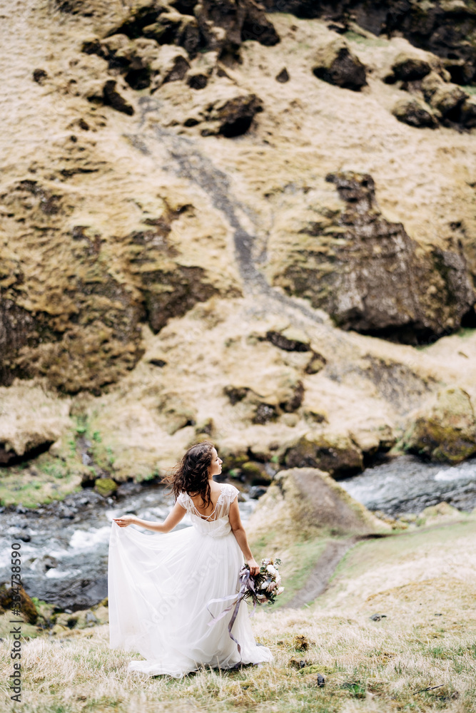 A bride in a white dress and a bouquet of flowers in her hands walks along the banks of a mountain river, near Kvernufoss waterfall in southern Iceland.