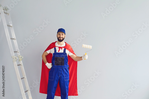 Happy home painter worker engaged in house renovation, holding roller brush, wearing superhero costume near grey wall