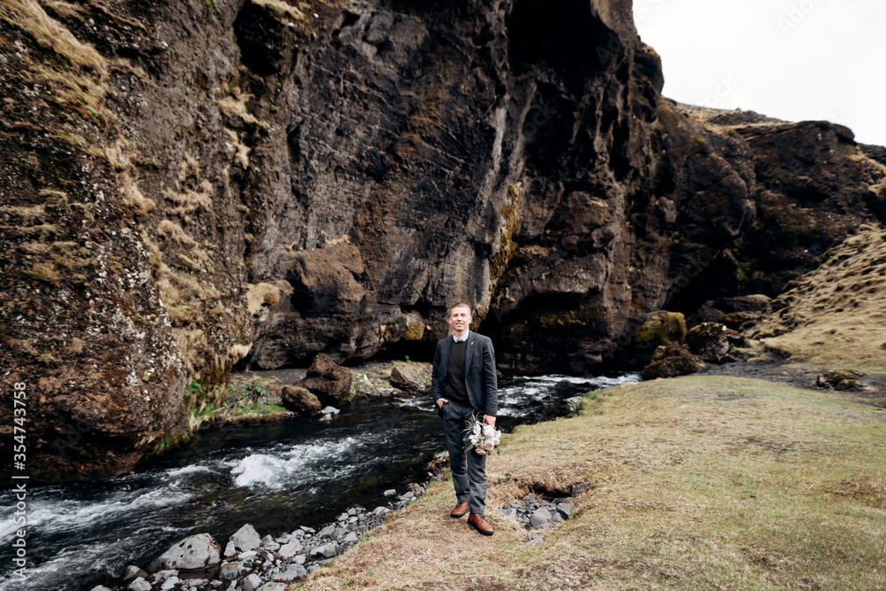 Destination Iceland wedding. A groom with a brides bouquet in hands is standing near a mountain river and a cliff. The right hand in the pocket of the trousers.