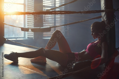 Young slim woman sitting at floor of boxing ring and relaxing in sunset lightning after training. Full length portrait