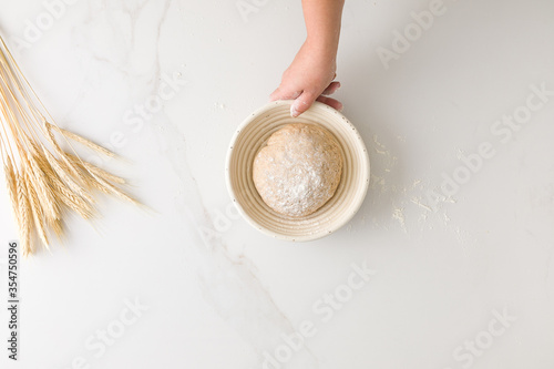 Top view of female hand holding a resting bread dough in a bread bowl in a marble table with wheat and flour with space for text
