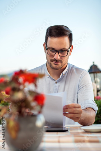 Obraz na płótnie Focused young Caucasian businessman looking over sales reports and documents while enjoying in his coffee break in riverside restaurant w salonie