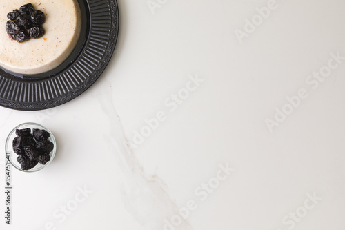Top view of a coconut pudding with plums in a black plate in a marble table with a glass bowl of plums and space for text