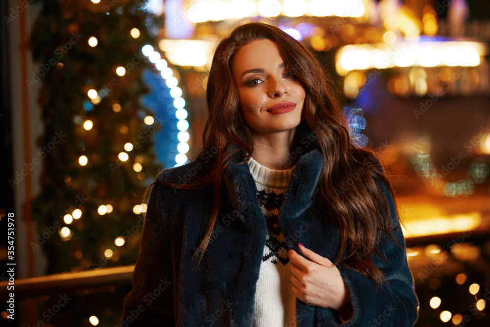 Closeup face portrait of young beautiful caucasian brunette woman with long wavy hair in bue coat looking at you. Christmas decorated city street at background