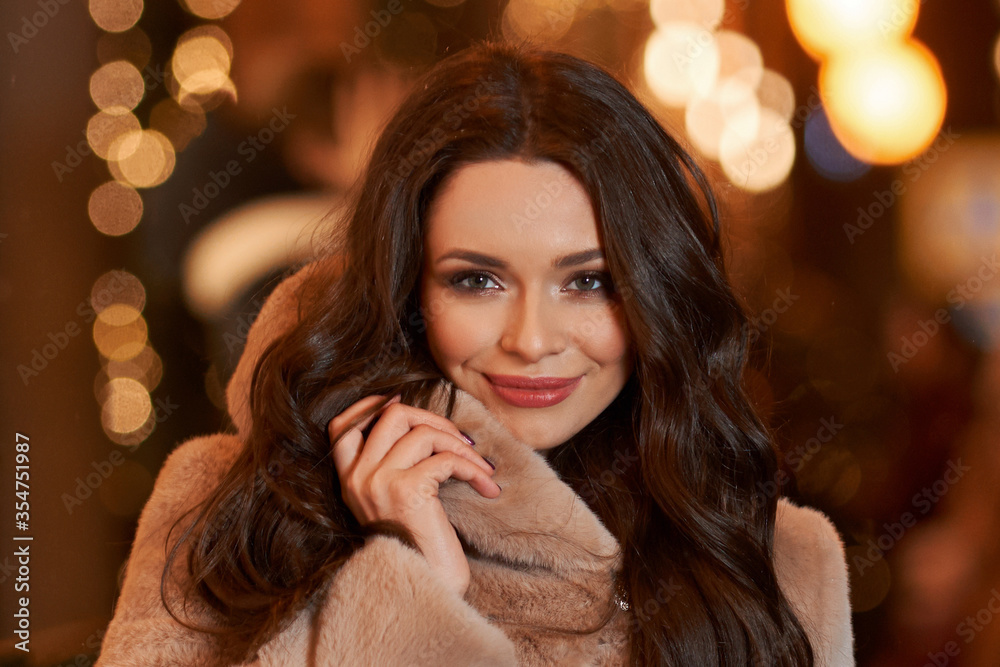 Closeup face portrait of young beautiful caucasian brunette woman with long wavy hair in pink coat looking at you. Christmas decorated city street at background