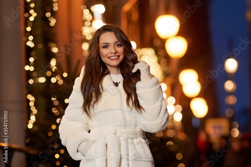 Closeup face portrait of young beautiful caucasian brunette woman with long wavy hair in white coat looking at you. Christmas decorated city street at background