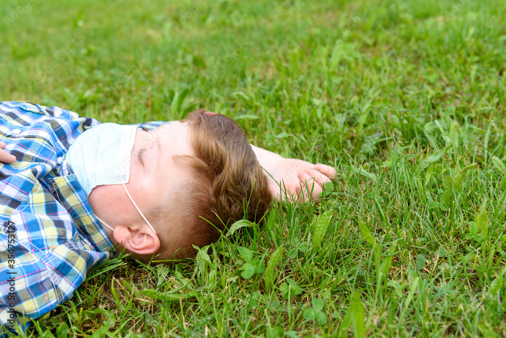 Portrait of a young boy with surgical mask lying on the grass in the park. Close-up of a little boy in a plaid shirt relaxing on the grass. Portrait of a young boy with surgical mask lying on the grass in the park. Close-up of a little boy in a plaid shirt relaxing on the grass.