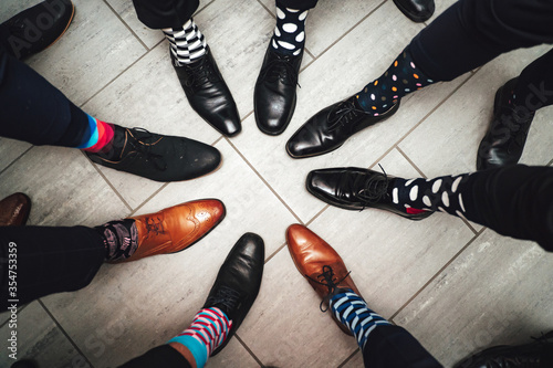 Men feet in shoes and funny colorful socks in circle composition. Top view of 8 legs wearing formal leather shoes on grey tiled floor.