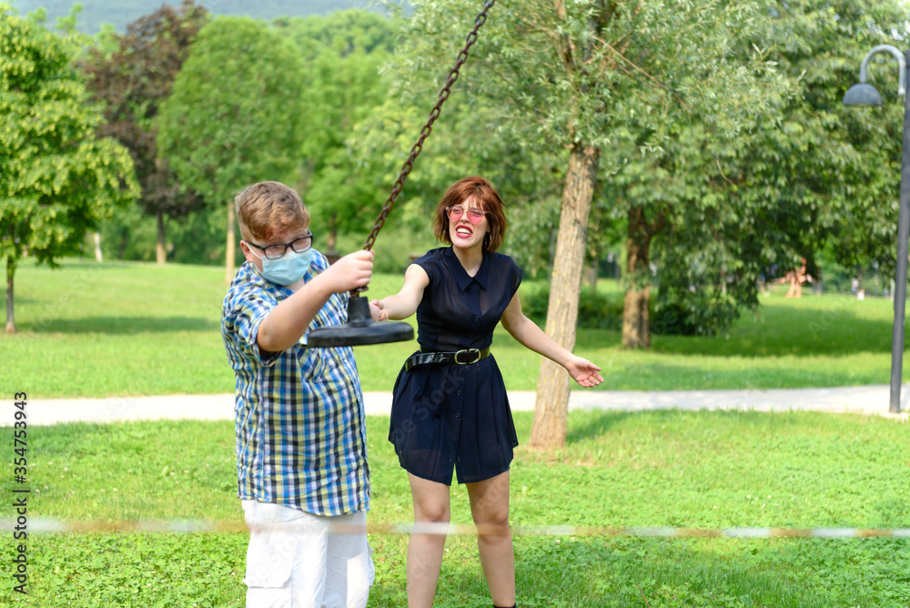 Obraz premium Boy wearing eyeglasses and surgical mask dressed in a plaid shirt plays with an adult girl in front of a red and white striped barrier tape around the playground. 