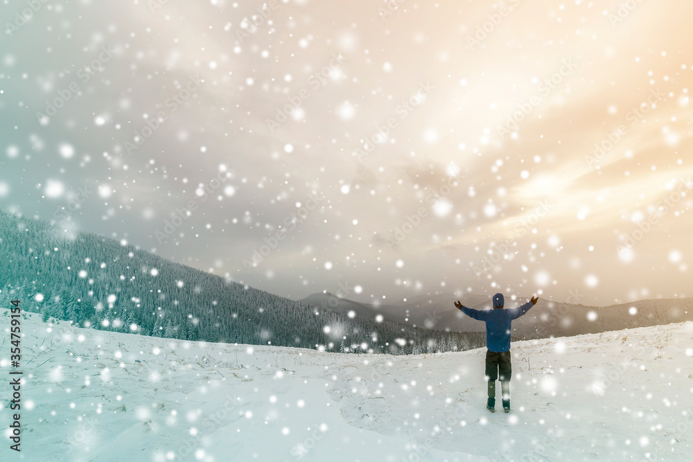 Back view of tourist hiker in warm clothing with backpack standing with raised arms on clearing covered with snow on spruce forest mountain and cloudy sky copy space background. Back view of tourist hiker in warm clothing with backpack standing with raised arms on clearing covered with snow on spruce forest mountain and cloudy sky copy space background.