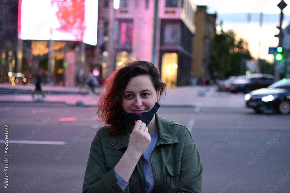 Fototapeta premium Smiling young woman removes the black protective mask on the street of a big city. Anti coronavirus, virus and flu concept. Back to normal life after pandemic. Quarantine is over. Urban evening scene.