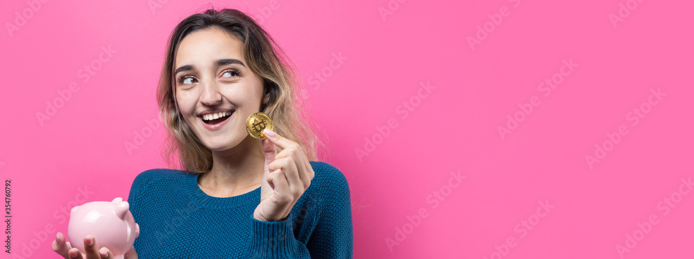 Girl puts in the piggy bank physical Bitcoin.Young girl over pink background holding piggy bank and rejoices gesturing. The concept of reliability of cash investments and insurance. Girl puts in the piggy bank physical Bitcoin.Young girl over pink background holding piggy bank and rejoices gesturing. The concept of reliability of cash investments and insurance.