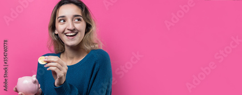 Girl puts in the piggy bank physical Bitcoin.Young girl over pink background holding piggy bank and rejoices gesturing. The concept of reliability of cash investments and insurance.
