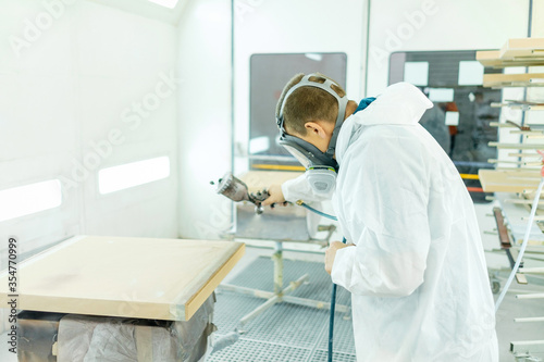 Painter with safety mask painting a wooden furniture with spray gun. Worker painting furniture details at the spray booth.