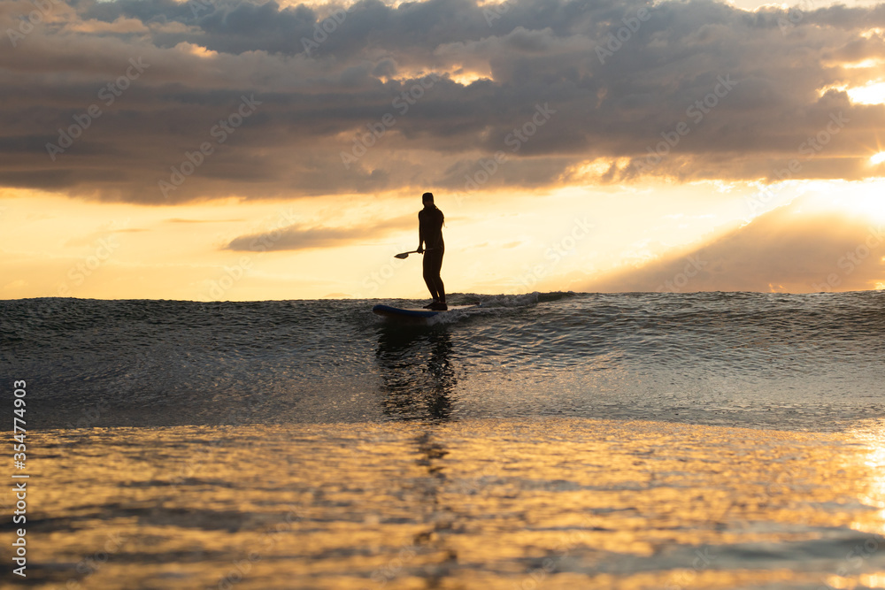 Stand Up Paddle Boarding In Japan at Sunrise and Sunset a solo rider keeping fit & healthy on the Pacific Ocean in a black wetsuit, also catching some large waves, The ocean is blue with a nice sky.
