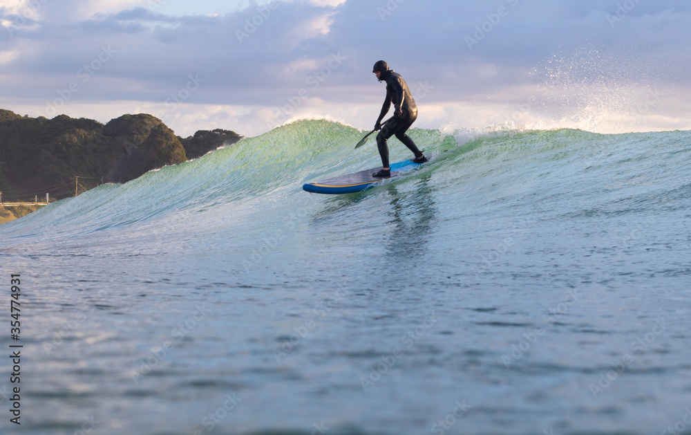 Naklejka premium Stand Up Paddle Boarding In Japan at Sunrise and Sunset a solo rider keeping fit & healthy on the Pacific Ocean in a black wetsuit, also catching some large waves, The ocean is blue with a nice sky.