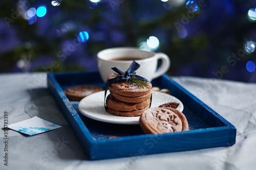 Creative Christmas homemade biscuits with blue ribbon and twig of fir on a white plate on blue background. A cup of tea and blue lights on a background. Side view