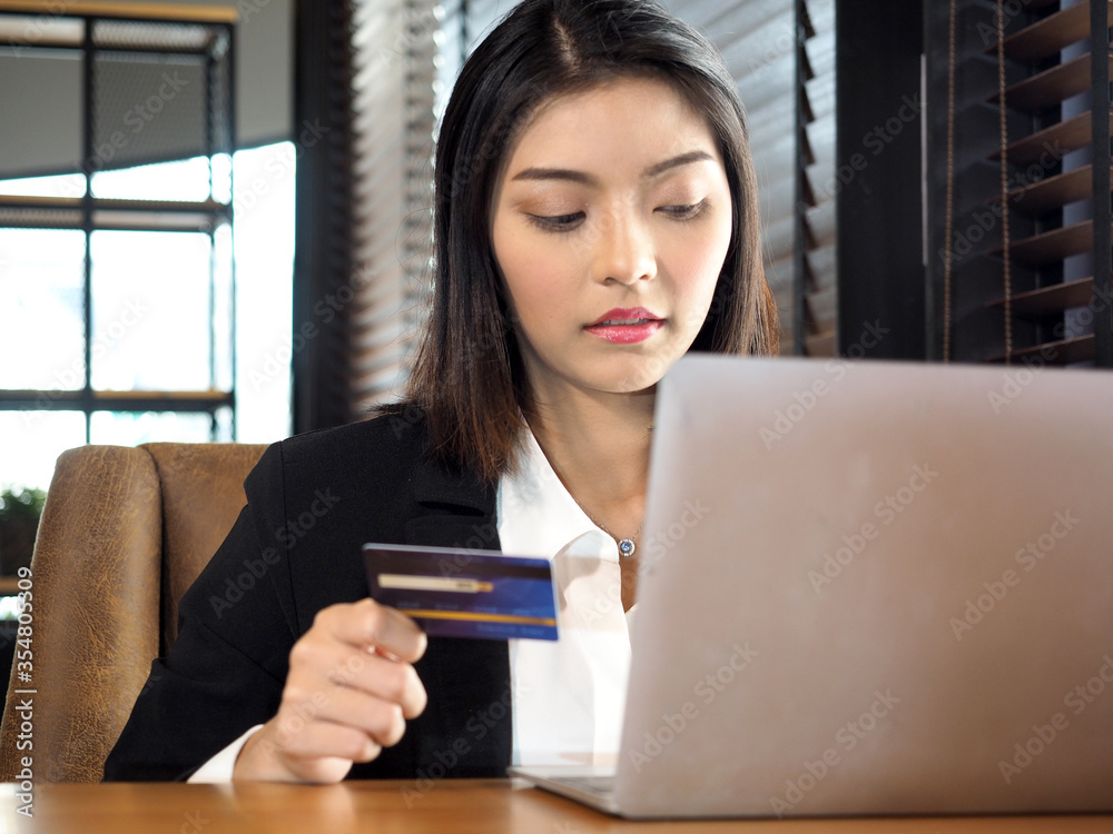 Fototapeta premium Asian business woman in suit using credit card shopping online with computer laptop in coffee shop cafe.Young female shopping online and paying with a credit card. 